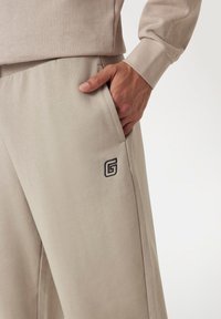 Bežine trenirke s bočnom džepom i izvezenim logom. Mekani materijal i opušteni kroj s lagano suženim nogavicama.