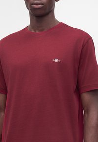 Homme portant un t-shirt rouge uni avec un petit logo blanc brodé sur la poitrine gauche sur un fond uni.