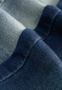 Tissu en denim dans des nuances de bleu variées, présentant une surface texturée avec une finition lisse et de subtils dégradés de couleur, replié sur lui-même.