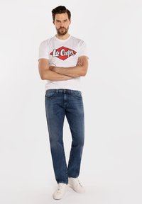 Mężczyzna z brodą i wąsami stoi ze skrzyżowanymi ramionami, ubrany w białą koszulkę Lee Cooper, niebieskie dżinsy i białe trampki na białym tle.