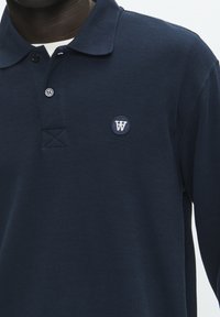 Jūras zilas krāsas polo krekls, izgatavots no teksturēta auduma, ar divu pogu stilu un apaļu logotipu kreisajā krūšu daļā.