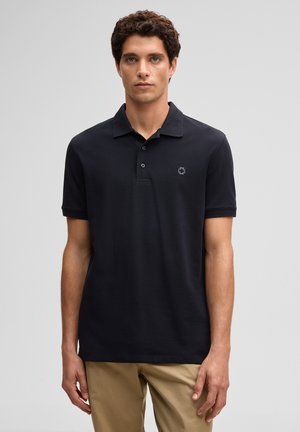 Junger Mann mit lockigem braunem Haar, der ein schwarzes Polohemd mit kurzen Ärmeln und beige Hose trägt, vor einem schlichten grauen Hintergrund stehend.