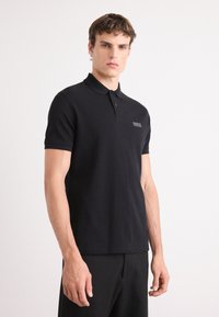 Polo noir en tissu coton doux. Manches courtes, col classique, patte de boutonnage à trois boutons, avec un discret logo brodé sur la poitrine.