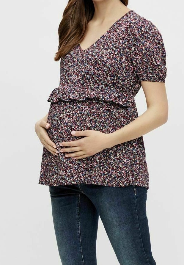 Maglietta da maternità con stampa floreale, scollo a V e maniche corte, caratterizzata da un orlo a balze. Indossata con jeans scuri, esalta la silhouette di una donna incinta.