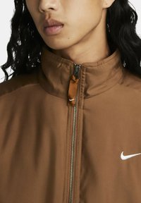 Hnědá bundu na zip s vysokým límcem, vyrobená z hladkého materiálu. Obsahuje stříbrný zip a bílé logo Nike na levé straně hrudi.