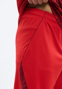 Κοντινό πλάνο ενός ατόμου που φορά κόκκινα αθλητικά σορτς με σκουρόχρωμες κόκκινες πλευρικές λωρίδες και ελαστική μέση, με φόντο μια απλή επιφάνεια.