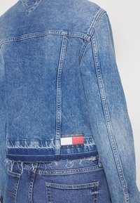 Džínová bunda ve středně modrém odstínu s roztrhaným lemem, tvarovaným výstřihem na zádech do V a s označením značky na levé straně v pase.