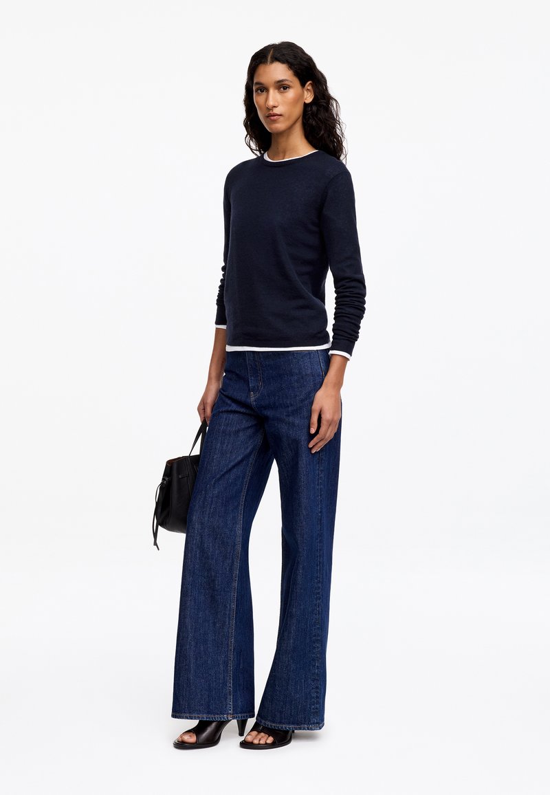 Femme aux longs cheveux foncés portant un pull noir par-dessus un top blanc, un jean bleu foncé à jambes larges, des sandales à talons noires, tenant un sac à main noir.