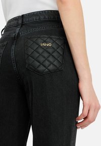 Ingrandimento di jeans neri con una tasca posteriore in pelle trapuntata con il logo dorato LIU JO sul lato destro, indossati con una maglietta bianca.