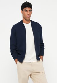 Námořnická bomber bunda na zip vyrobená z pleteného materiálu s žebrovanými manžetami a lemem, na levém prsou má malé logo.