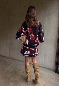 Γυναίκα με κυματιστά καστανά μαλλιά, φοράει μαύρο πουλόβερ με σχέδια και φούστα, κρατώντας μπεζ τσάντα χειρός, στέκεται με μπεζ μπότες μέχρι το γόνατο μπροστά από τσιμεντένιο τοίχο.