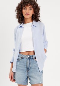 Chemise boutonnée rayée bleu clair et blanche sur un crop top blanc, associée à un short en denim taille haute déchiré.