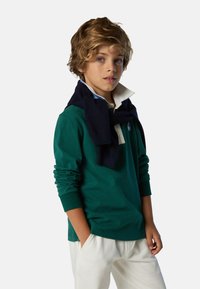 Polo verde de manga larga con un suéter azul marino sobre los hombros, combinado con pantalones blancos, con cuello doblado y detalle de logo.