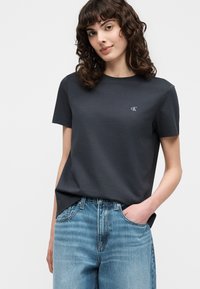 Νεαρή γυναίκα με σγουρά μαλλιά που φοράει ένα απλό σκούρο γκρι T-shirt CK και ανοιχτό μπλε τζιν, χέρια στις τσέπες, σε λευκό φόντο.