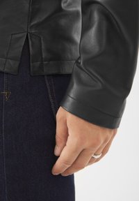 Musta nahkatakki, jossa on sileä pinta ja istuvat hihat, ja näkyvissä on käsi, joka lepää tumman denimkankaan lähellä.