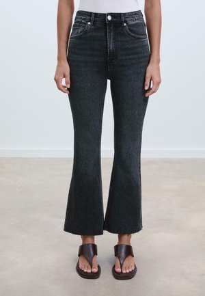 Personne portant un jean flare court taille haute gris foncé avec ourlet brut et des sandales ouvertes marron, debout sur un sol clair.