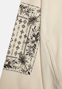 Felpa crema con tessuto testurizzato. Presenta una stampa floreale e geometrica nera sulla manica, inclusi palme e un bordo decorativo.