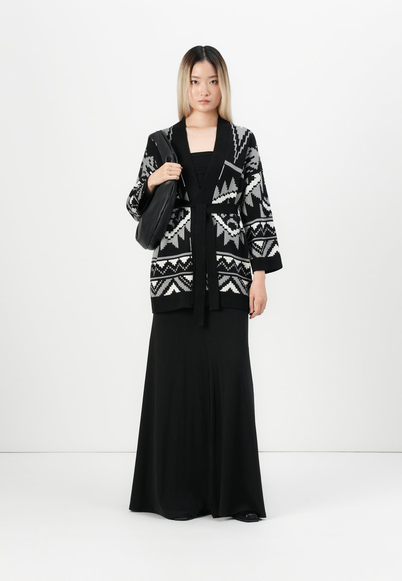 Cardigan a righe in bianco e nero con cintura, indossato sopra un vestito aderente nero. L'outfit comprende una borsa e maniche lunghe, posizionato su uno sfondo semplice.