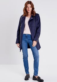 Marineblauer Kunstsuede-Mantel mit asymmetrischem Saum, silbernen Reißverschlüssen und seitlichen Reißverschlusstaschen, kombiniert mit einem leichten Mesh-Oberteil und blauen Jeans.