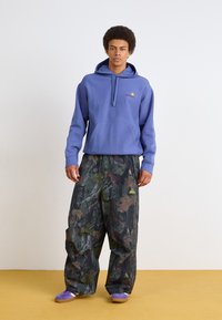 Purple hoodie, camouflage wide-leg pants with green and brown leaf patterns, and purple slip-on shoes. Features logo accent on hoodie and pants.