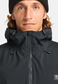 Schwarze Zip-Jacke mit hohem Kragen, Kordelzugverstellung und einem Logo-Patch. Glatte Textur mit mattem Finish und verdeckten Reißverschlüssen.