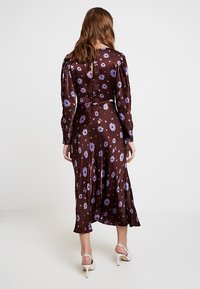 Brun klänning med blommönster, långa ärmar, en svävande nederdel och en knäppt baksida. Har lila blommor och en mjuk tygtextur.