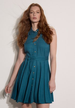 Femme aux cheveux roux portant une robe sans manches bleu sarcelle à boutons sur le devant, avec col et ceinture à la taille, debout devant un fond clair uni.