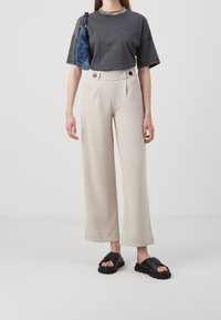 T-shirt grigio oversize, pantaloni beige a gamba larga con pieghe e sandali neri a scivolo, con una borsa a fantasia blu e una collana in argento.