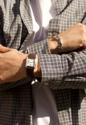 Person in plaid jacket with arms crossed, wearing a rectangular watch with brown leather strap and a silver wavy bracelet.