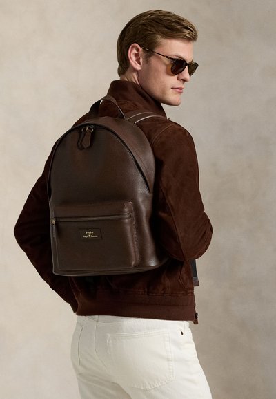 Mann mit Sonnenbrille, dunkelbrauner Jacke und weißen Hosen trägt einen dunkelbraunen Lederrucksack mit Frontreißverschlusstasche.