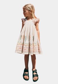 Helles beige Baumwollkleid mit gestickten farbenfrohen Mustern, Rüschenärmeln und einem ausgestellten Rock. Schwarze Sandalen mit Schnallenriemen.