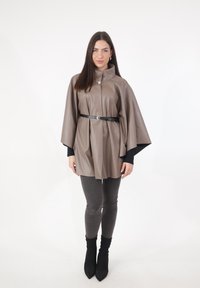 Brauner Poncho aus Kunstleder mit hohem Kragen, in der Taille mit einem schwarzen Gürtel gerafft. Trägt man über schwarzen Leggings und spitzen Stiefeletten.