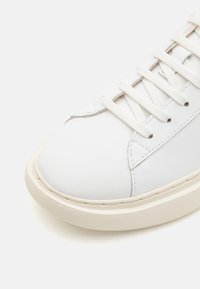 Gros plan sur l'avant d'une basket en cuir blanc avec des lacets blancs et une semelle en caoutchouc beige sur fond blanc.