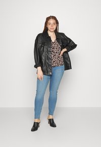 Casaco oversized de couro preto, blusa com padrão de leopardo, jeans skinny azul claro e mules pretos com a ponta aberta. Fundo simples, cor neutra.