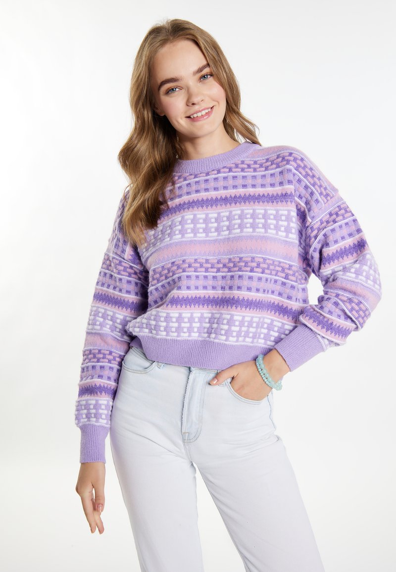 Cardigan viola con motivi a tonalità variabili, caratterizzato da disegni geometrici e un colletto rotondo a coste, abbinato a pantaloni di jeans chiari.