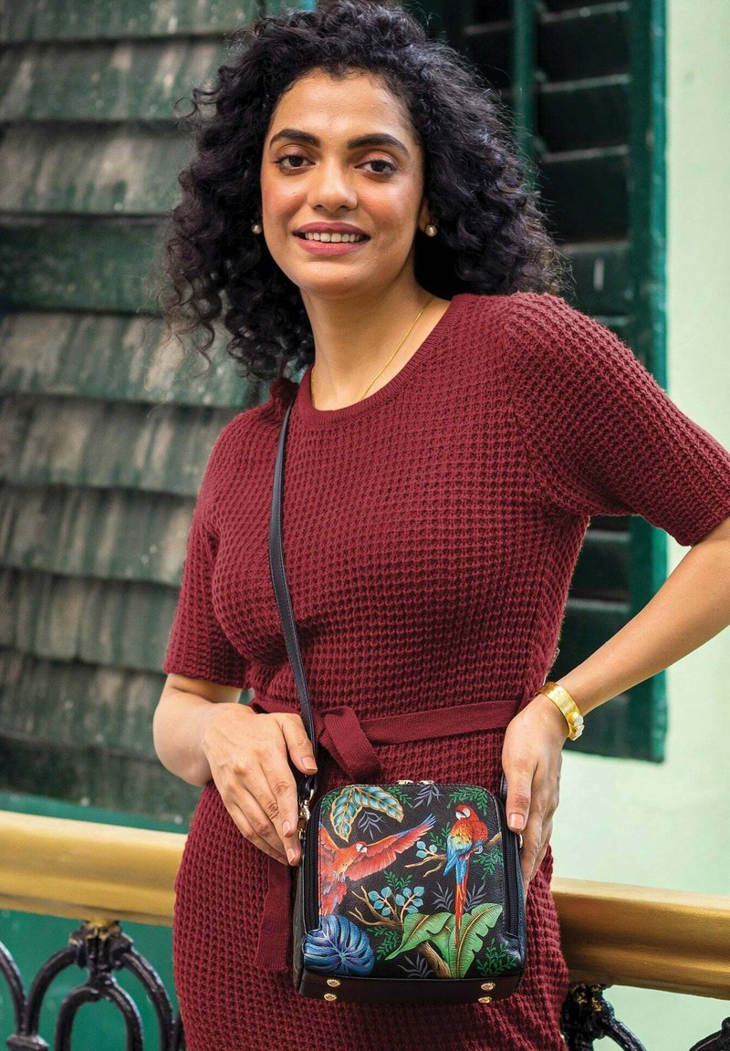 Zwarte schoudertas met een levendig ontwerp van tropische vogels. Bevat kleurrijke loofbladeren en gouden hardware accenten. Geweven rode jurk op de achtergrond.