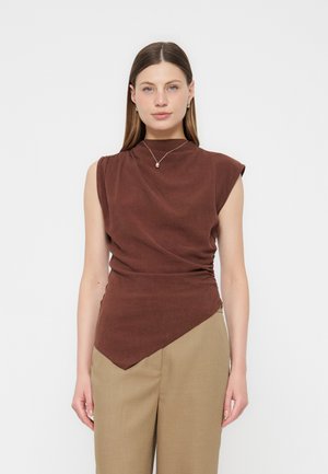 Γυναίκα με ίσια καστανά μαλλιά που φοράει αμάνικο καφέ τοπ με ασύμμετρη λήξη και μπεζ παντελόνι με ψηλή μέση, στέκεται μπροστά σε λευκό φόντο.