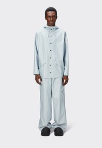 Chaqueta impermeable de color azul claro con capucha, botones a presión y bolsillos delanteros, combinada con pantalones holgados a juego y zapatos negros.