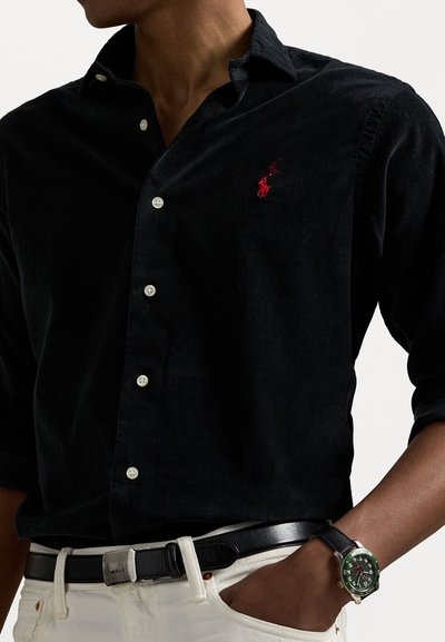 Black corduroy button-up shirt with a small red logo, white buttons, and rolled sleeves, paired with black belt and silver-green watch.
