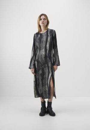 Robe longue noire avec des motifs abstraits gris, dotée de manches longues, de fentes latérales et d'une texture lisse. Associée à des bottines noires.