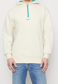 Sweat à capuche crème avec accents turquoise et fermeture à demi-fermeture éclair, doté d'une poche avant et d'un logo brodé sur la poitrine. Tissu texturé.