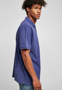 T-shirt polo violet à manches courtes, avec fentes latérales et col classique, associé à un jean bleu clair, présenté de profil.