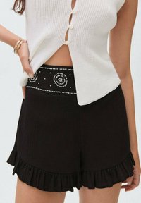 Black shorts with a textured fabric, featuring a ruffled hem and a decorative waistband with white bead patterns. White knit top partially visible.
