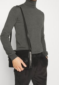 Schwarze Leder-Umhängetasche mit Klappe, verstellbarem Schultergurt und Frontschnallendetail. Glatte Textur und elegantes Design.