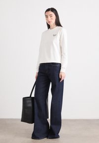 Ung kvinne står med en svart tote bag, iført en hvit hettegenser og mørke vide jeans, satt mot en minimalistisk bakgrunn.