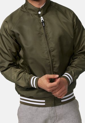 Man wearing an olive green satin bomber jacket with white striped ribbed cuffs and waistband, and light gray pants.