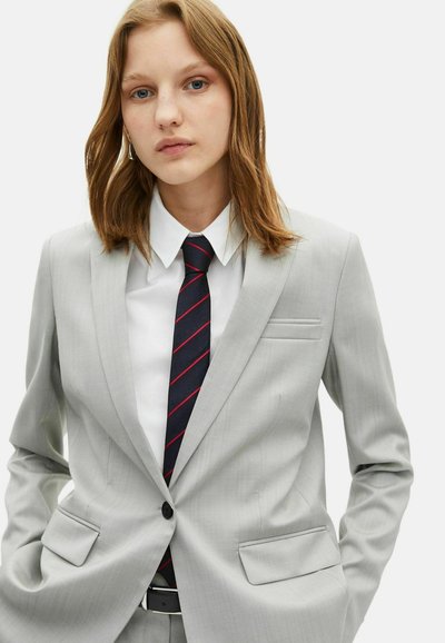Blazer grigio chiaro su misura, chiusura a singolo bottone, rever a lancia, due tasche frontali, indossato sopra una camicia bianca e una cravatta scura con strisce rosse.