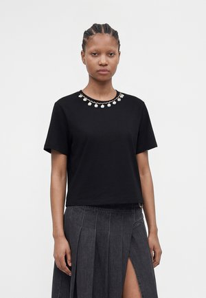 Mujer con cabello trenzado que lleva una camiseta negra de manga corta con detalle de collar de conchas y una falda oscura plisada con una abertura alta en el muslo.