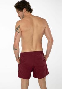 Un maillot de bain bordeaux avec une taille élastique, deux poches latérales et une petite poche arrière, montré de dos sur un modèle à la peau nue.