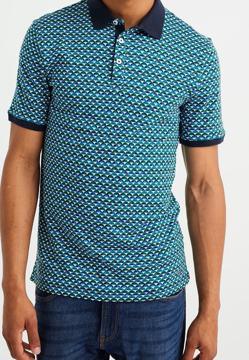 Homme portant un polo à manches courtes avec un motif géométrique bleu, vert et blanc, avec un col et des poignets foncés, associé à un jean bleu foncé.